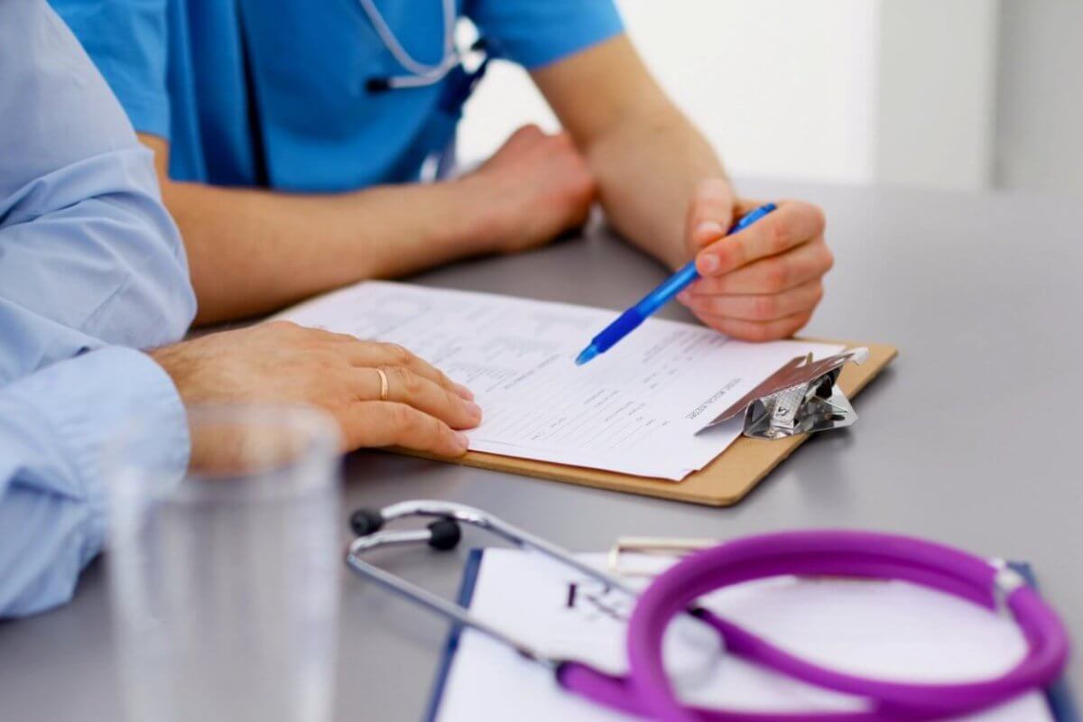 A close-up image of two family medicine doctors in Metro Manila reviewing a document on a clipboard. One person is holding a blue pen and pointing at the paper. The table has a stethoscope and additional papers. Both individuals are wearing medical scrubs, and there's a glass of water in the foreground.