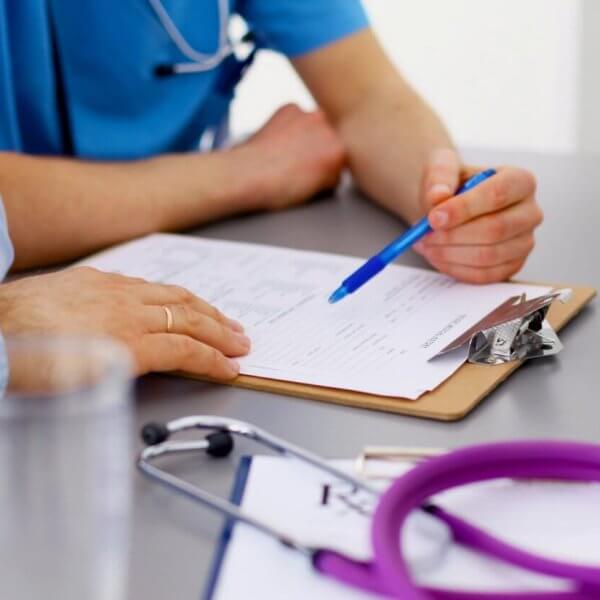 A close-up image of two family medicine doctors in Metro Manila reviewing a document on a clipboard. One person is holding a blue pen and pointing at the paper. The table has a stethoscope and additional papers. Both individuals are wearing medical scrubs, and there's a glass of water in the foreground.