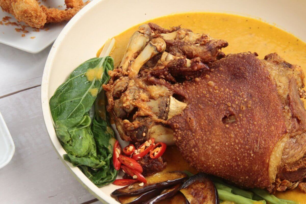 A must-try dish featuring crispy, deep-fried pork knuckle sits in a yellow curry sauce, accompanied by vegetables like leafy greens, eggplant slices, and red chili peppers. In the background, breaded shrimp and a small dish of dipping sauce are partially visible on white plates—an experience you can't miss at Filipino restaurants.
