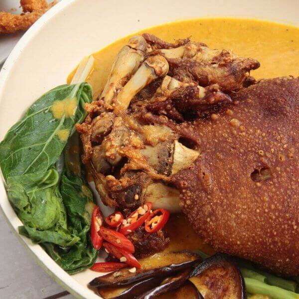 A must-try dish featuring crispy, deep-fried pork knuckle sits in a yellow curry sauce, accompanied by vegetables like leafy greens, eggplant slices, and red chili peppers. In the background, breaded shrimp and a small dish of dipping sauce are partially visible on white plates—an experience you can't miss at Filipino restaurants.