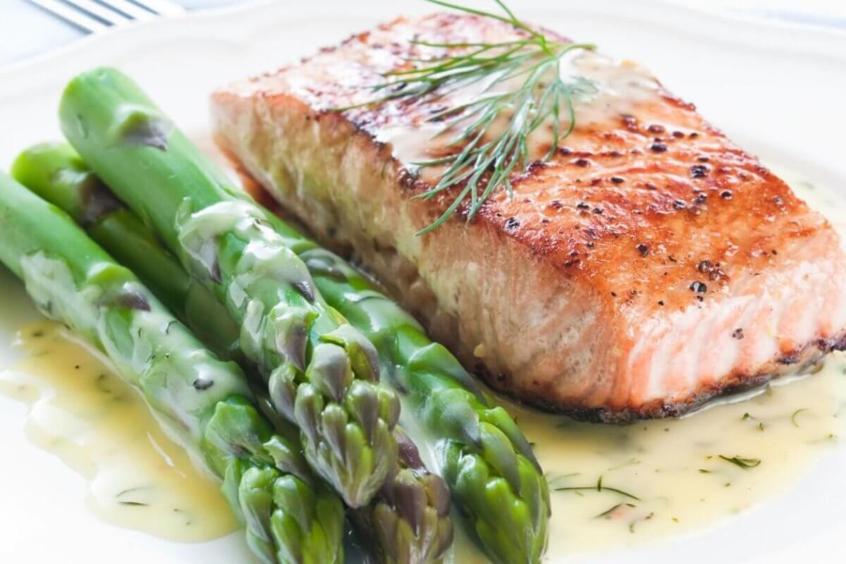 A plate of food featuring a grilled salmon fillet garnished with fresh dill, accompanied by several spears of steamed asparagus. The dish is drizzled with a creamy dill sauce, showcasing the best Greenhills dining experience. The plate sits on a white tablecloth with a fork partially visible in the background.