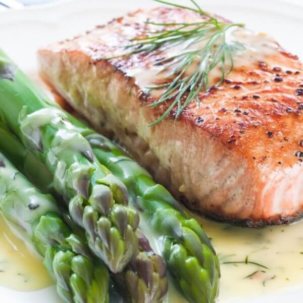 A plate of food featuring a grilled salmon fillet garnished with fresh dill, accompanied by several spears of steamed asparagus. The dish is drizzled with a creamy dill sauce, showcasing the best Greenhills dining experience. The plate sits on a white tablecloth with a fork partially visible in the background.
