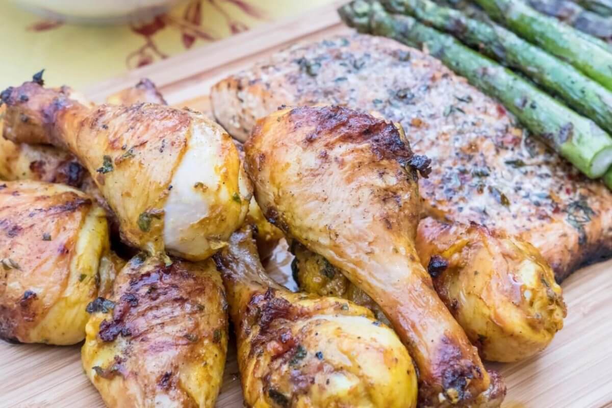 Grilled chicken drumsticks, seasoned and charred, are arranged in the foreground on a wooden board, alongside a piece of herb-topped inihaw fish. To the side, there's a row of grilled asparagus spears, adding a vibrant green contrast to this delightful dish reminiscent of popular hotspots in Metro Manila.