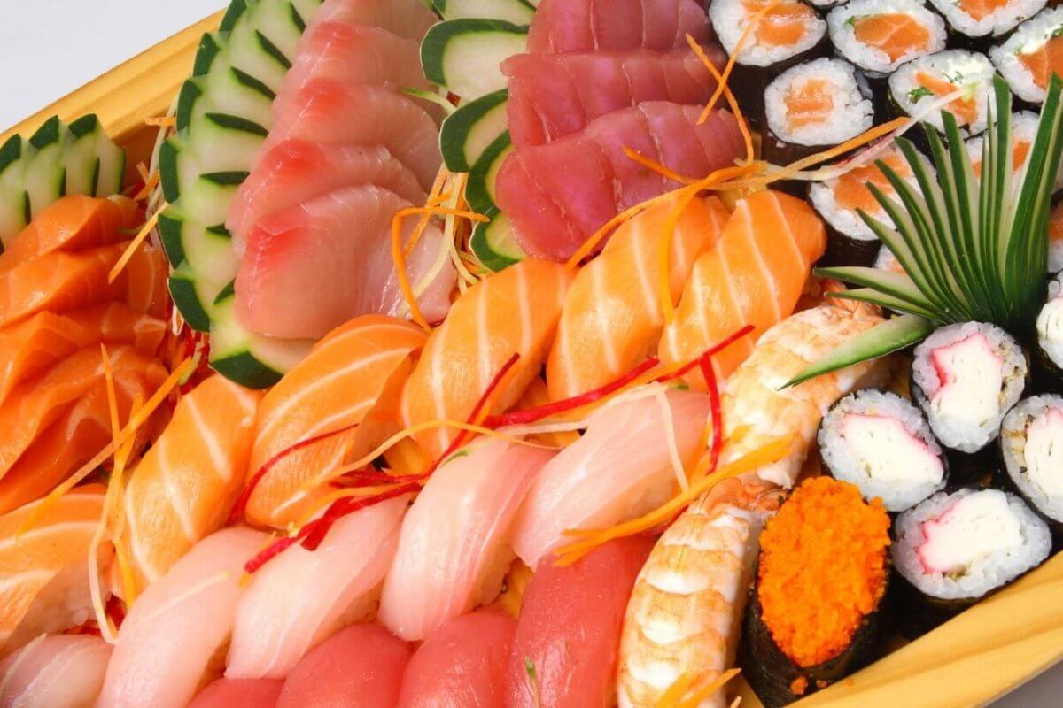 A large wooden platter filled with an assortment of sushi and sashimi, reminiscent of offerings found in Japanese grocery stores across Metro Manila. The platter includes various kinds of fish like salmon, tuna, and white fish, garnished with thin vegetable slices and shredded carrots. There are also sushi rolls, some topped with orange roe, and garnished with a clump of green grass.