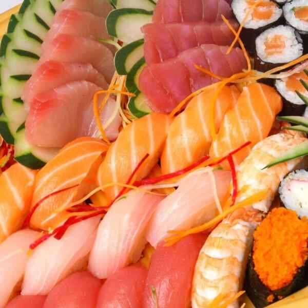 A large wooden platter filled with an assortment of sushi and sashimi, reminiscent of offerings found in Japanese grocery stores across Metro Manila. The platter includes various kinds of fish like salmon, tuna, and white fish, garnished with thin vegetable slices and shredded carrots. There are also sushi rolls, some topped with orange roe, and garnished with a clump of green grass.