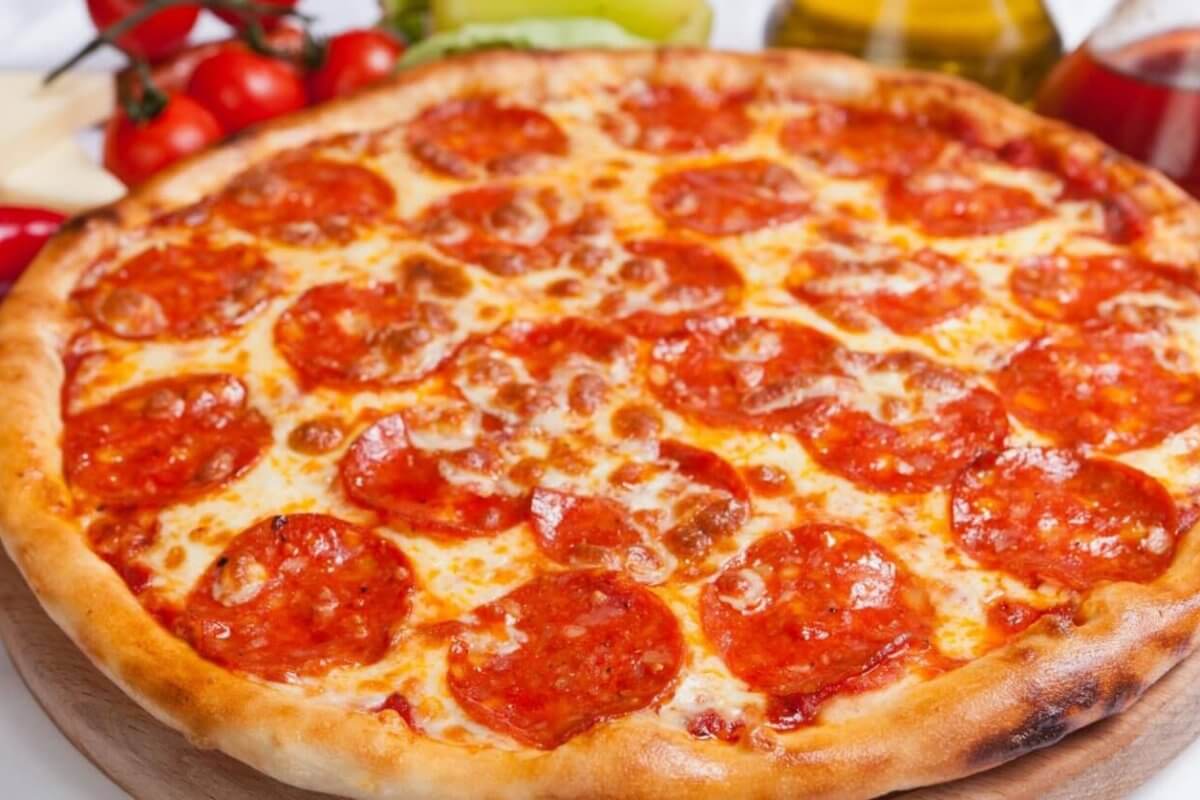 A freshly baked pepperoni pizza with a golden-brown crust is displayed on a wooden board. The pizza, reminiscent of what you might find at top restaurants, is topped with melted cheese and an uneven spread of pepperoni slices. Ingredients like tomatoes, peppers, cheese, and oil bottles are blurred in the background.