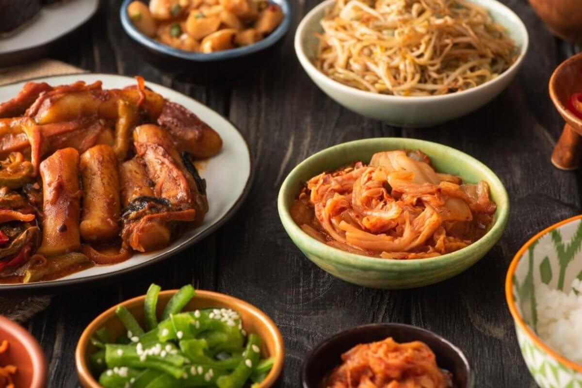 A variety of Korean dishes are displayed on a dark wooden table. The spread, which showcases the best of Korean cuisine, includes a plate of stir-fried meat and vegetables, a bowl of sauteed bean sprouts, kimchi from one of Metro Manila’s top Korean groceries, sliced red chili peppers, green beans with sesame seeds, white rice, and small dishes of other sides.