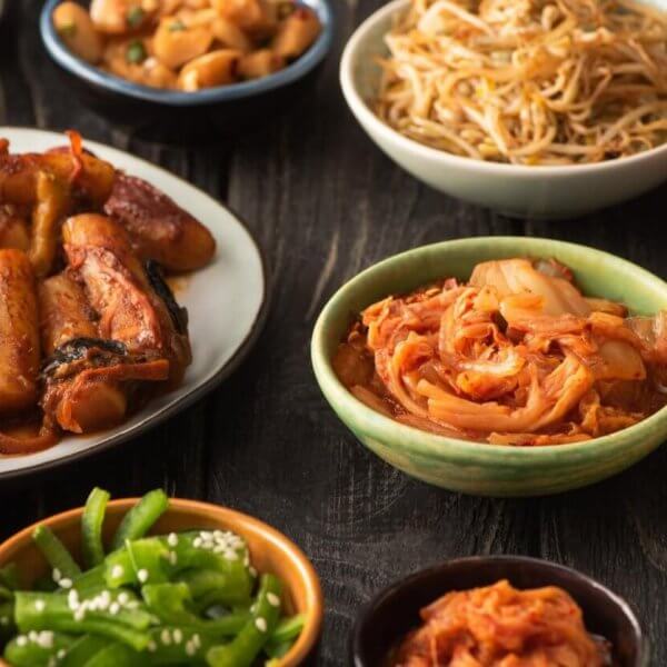 A variety of Korean dishes are displayed on a dark wooden table. The spread, which showcases the best of Korean cuisine, includes a plate of stir-fried meat and vegetables, a bowl of sauteed bean sprouts, kimchi from one of Metro Manila’s top Korean groceries, sliced red chili peppers, green beans with sesame seeds, white rice, and small dishes of other sides.