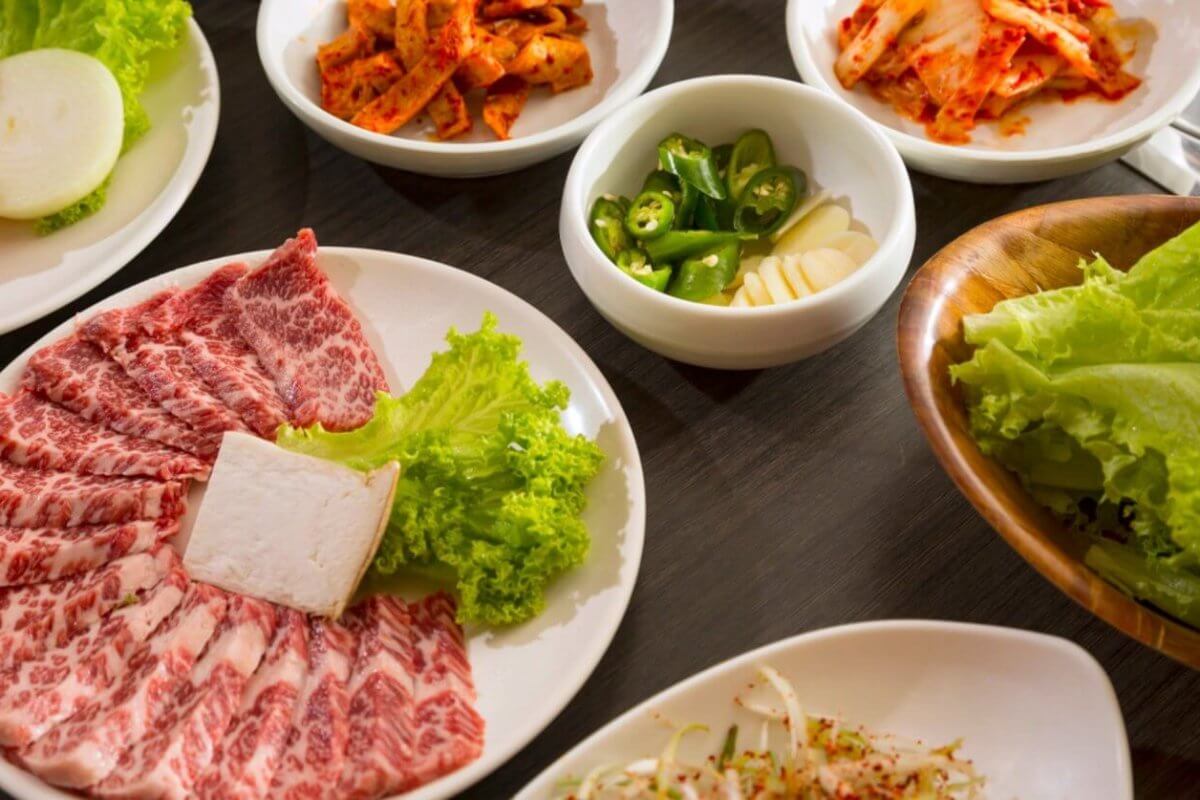 An array of Korean BBQ ingredients on a wooden table, featuring a plate of marbled beef slices with a square of tofu and lettuce, a platter of fresh lettuce leaves, and smaller bowls containing sliced onions, kimchi, green chili peppers, garlic cloves, and a mixed vegetable side dish—all part of the ultimate dining experience at top Korean restaurants in Metro Manila.