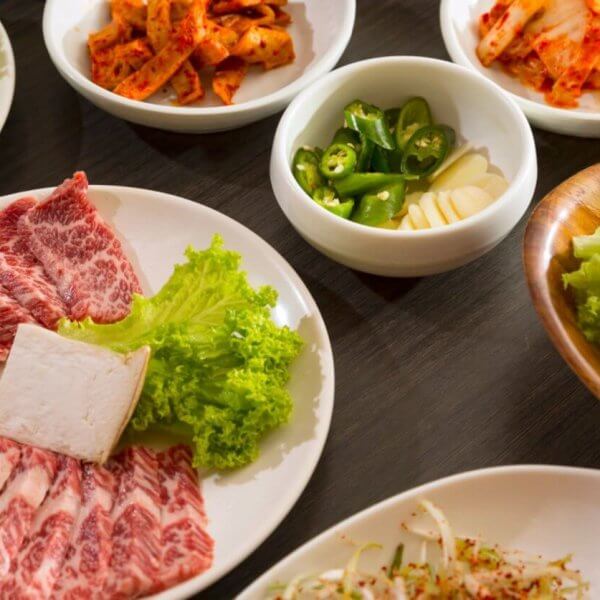 An array of Korean BBQ ingredients on a wooden table, featuring a plate of marbled beef slices with a square of tofu and lettuce, a platter of fresh lettuce leaves, and smaller bowls containing sliced onions, kimchi, green chili peppers, garlic cloves, and a mixed vegetable side dish—all part of the ultimate dining experience at top Korean restaurants in Metro Manila.