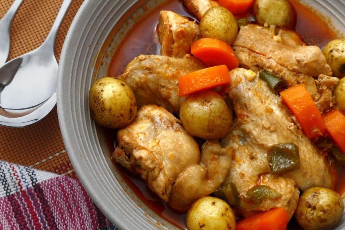 A savory dish of chicken stew is served in a grey bowl. The stew includes tender chicken pieces, whole baby potatoes, sliced carrots, and green bell pepper chunks, all immersed in a rich, red-colored broth. A fork and spoon are placed beside the bowl, resting on a colorful cloth napkin—an absolute must-try at Legaspi Village restaurants.