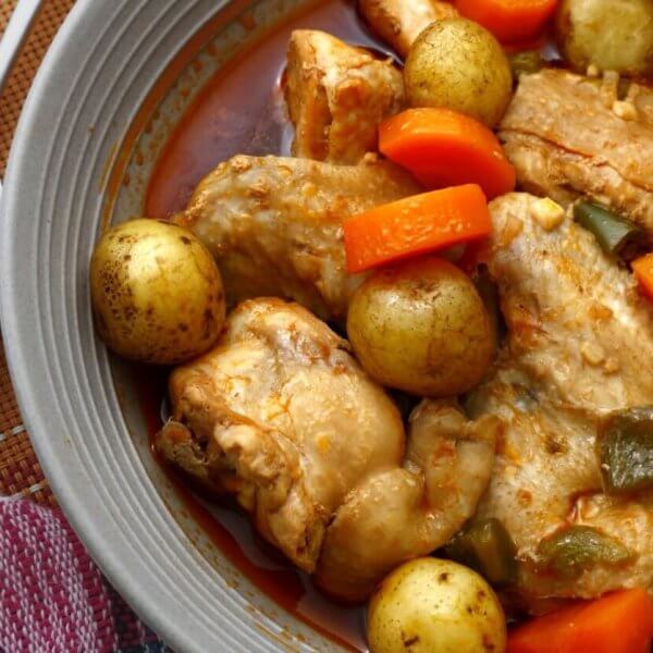 A savory dish of chicken stew is served in a grey bowl. The stew includes tender chicken pieces, whole baby potatoes, sliced carrots, and green bell pepper chunks, all immersed in a rich, red-colored broth. A fork and spoon are placed beside the bowl, resting on a colorful cloth napkin—an absolute must-try at Legaspi Village restaurants.