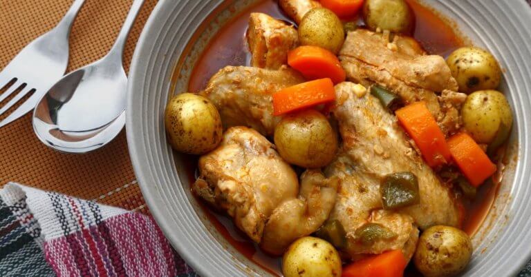 A savory dish of chicken stew is served in a grey bowl. The stew includes tender chicken pieces, whole baby potatoes, sliced carrots, and green bell pepper chunks, all immersed in a rich, red-colored broth. A fork and spoon are placed beside the bowl, resting on a colorful cloth napkin—an absolute must-try at Legaspi Village restaurants.