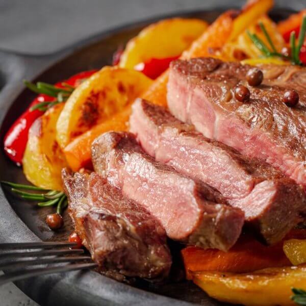 A juicy, medium-rare steak sliced into pieces sits on a black cast-iron skillet. The steak is garnished with whole peppercorns and fresh rosemary. Accompanying the steak are roasted potato wedges, red peppers, and a halved head of garlic. Nearby restaurants in Magallanes Village, Makati serve similar delights.