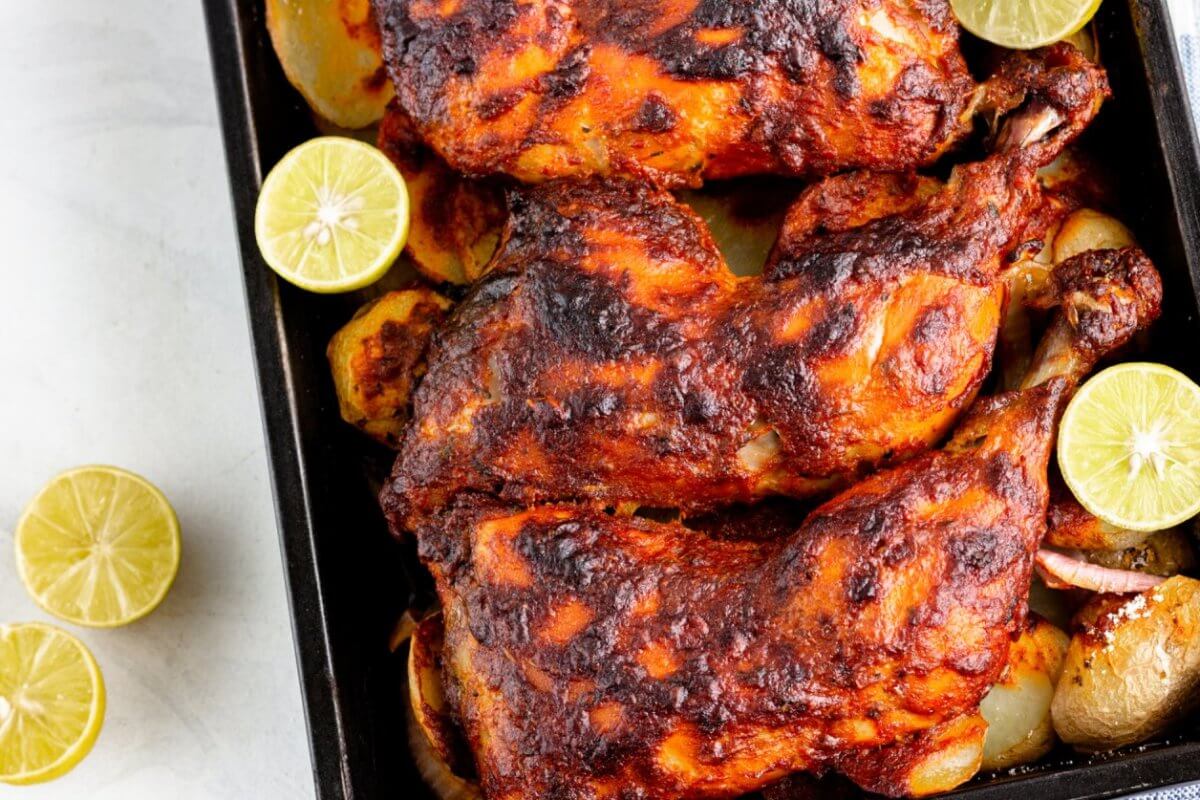 Three roasted chicken legs are placed on a baking tray, surrounded by roasted potato slices and garnished with halved limes. The chicken legs are marinated with a dark, crusty seasoning. Two halved limes rest on the white countertop next to the tray, reminiscent of dishes from restaurants along Marcos Highway.