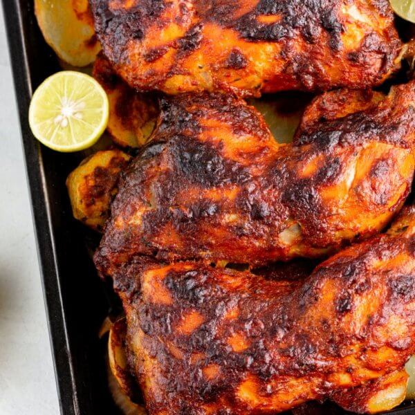 Three roasted chicken legs are placed on a baking tray, surrounded by roasted potato slices and garnished with halved limes. The chicken legs are marinated with a dark, crusty seasoning. Two halved limes rest on the white countertop next to the tray, reminiscent of dishes from restaurants along Marcos Highway.