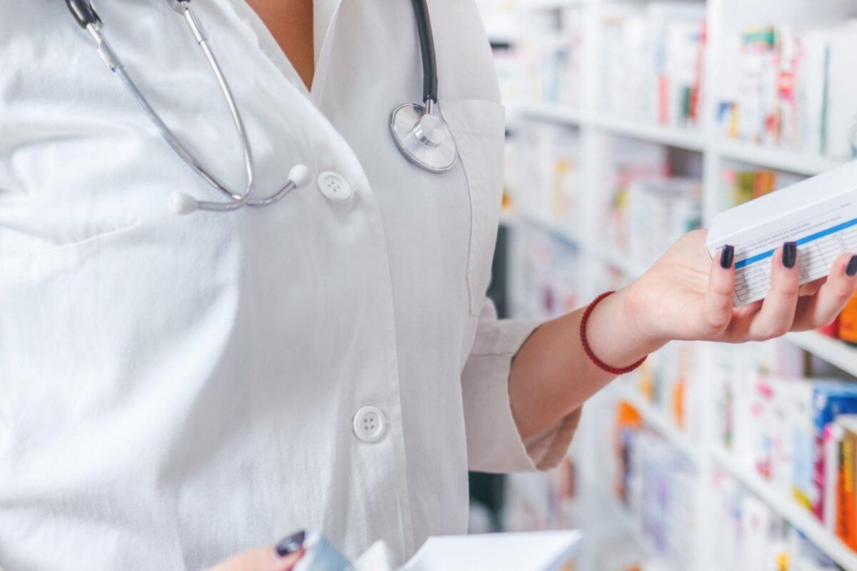 A pharmacist wearing a white coat and stethoscope is standing in a Mercury Branches pharmacy in Metro Manila. They are holding two boxes of medication while reading the label on one. Shelves filled with various medicine boxes are visible in the background.