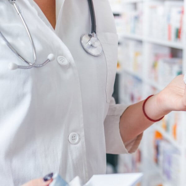 A pharmacist wearing a white coat and stethoscope is standing in a Mercury Branches pharmacy in Metro Manila. They are holding two boxes of medication while reading the label on one. Shelves filled with various medicine boxes are visible in the background.