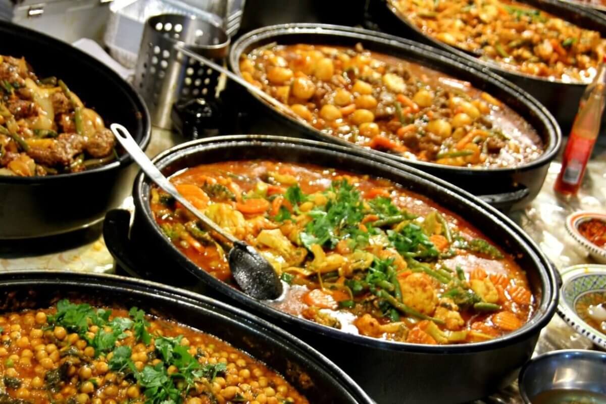 A hearty spread of various cooked dishes in large black pots is arranged on a table at the Eat-All-You-Can Buffet in SM Mall of Asia, featuring chickpea stew, meat and vegetable curry, and a tomato-based stew with herbs. Small bowls containing condiments and garnishes surround the main dishes, with serving spoons resting in the pots.