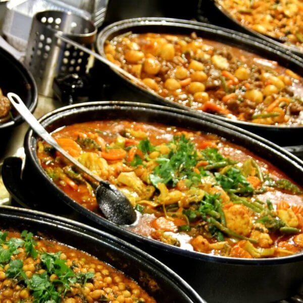 A hearty spread of various cooked dishes in large black pots is arranged on a table at the Eat-All-You-Can Buffet in SM Mall of Asia, featuring chickpea stew, meat and vegetable curry, and a tomato-based stew with herbs. Small bowls containing condiments and garnishes surround the main dishes, with serving spoons resting in the pots.
