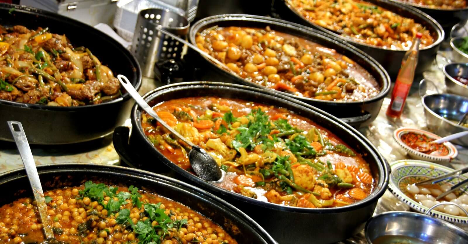 A hearty spread of various cooked dishes in large black pots is arranged on a table at the Eat-All-You-Can Buffet in SM Mall of Asia, featuring chickpea stew, meat and vegetable curry, and a tomato-based stew with herbs. Small bowls containing condiments and garnishes surround the main dishes, with serving spoons resting in the pots.