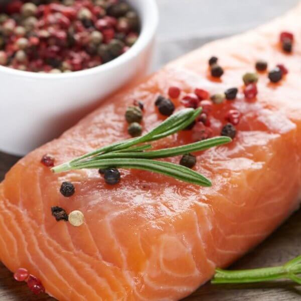 A raw salmon fillet is placed on a wooden surface, garnished with rosemary and an assortment of whole peppercorns. A small white bowl filled with mixed peppercorns is next to the salmon. Fresh cilantro sprigs lie beside the fillet, adding a touch of green to the presentation—a scene reminiscent of dishes at Molito Lifestyle Center's best restaurants.