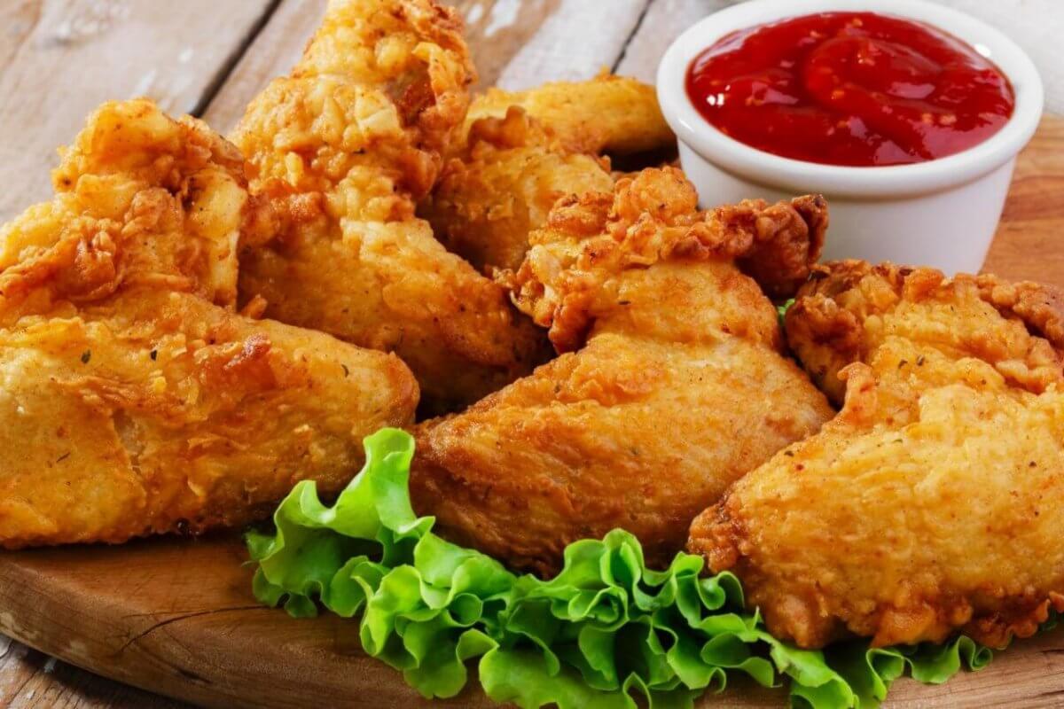 Close-up of crispy, golden-brown fried chicken pieces arranged on a wooden board, garnished with fresh green lettuce leaves at the bottom. A small white cup filled with red ketchup is placed behind the chicken. This must-try dish, found in Parañaque restaurants, sits against a rustic wooden background.