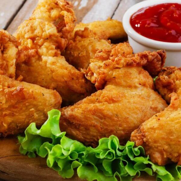 Close-up of crispy, golden-brown fried chicken pieces arranged on a wooden board, garnished with fresh green lettuce leaves at the bottom. A small white cup filled with red ketchup is placed behind the chicken. This must-try dish, found in Parañaque restaurants, sits against a rustic wooden background.