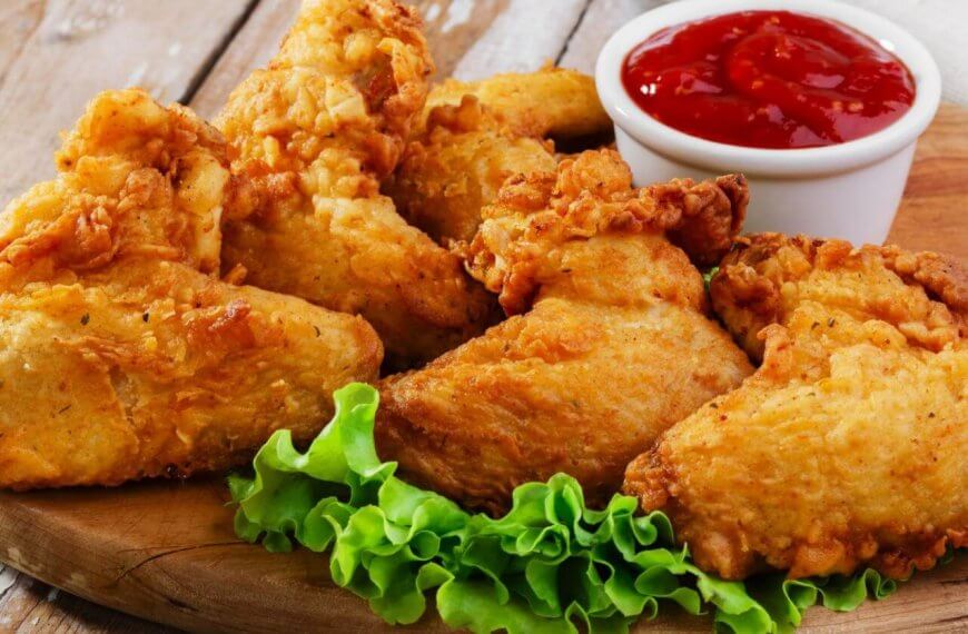 Close-up of crispy, golden-brown fried chicken pieces arranged on a wooden board, garnished with fresh green lettuce leaves at the bottom. A small white cup filled with red ketchup is placed behind the chicken. This must-try dish, found in Parañaque restaurants, sits against a rustic wooden background.
