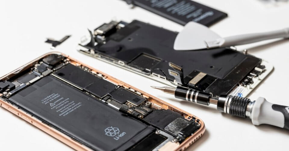 A disassembled smartphone with its internal components visible, including the battery and circuit board. Various repair tools such as screwdrivers and plastic spudgers are scattered around on a white surface. Another smartphone lies partially disassembled in the background, offering tips for buying a phone.