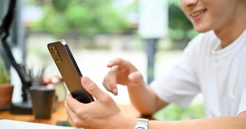 A person wearing a white shirt is smiling while holding their new phone in a dark-colored case. Seated at a wooden table adorned with small potted plants, they enjoy the natural light and greenery in the blurred background.