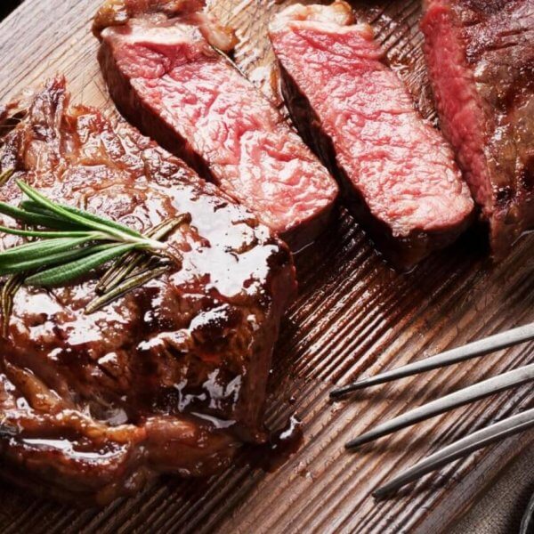 A wooden cutting board with a juicy, medium-rare steak is garnished with fresh rosemary and sliced into three pieces. A vintage fork rests on the board, beside a small bowl of colorful peppercorns. The neutral-toned cloth backdrop lends a charming vibe, reminiscent of delightful dining spots in Salcedo Village.