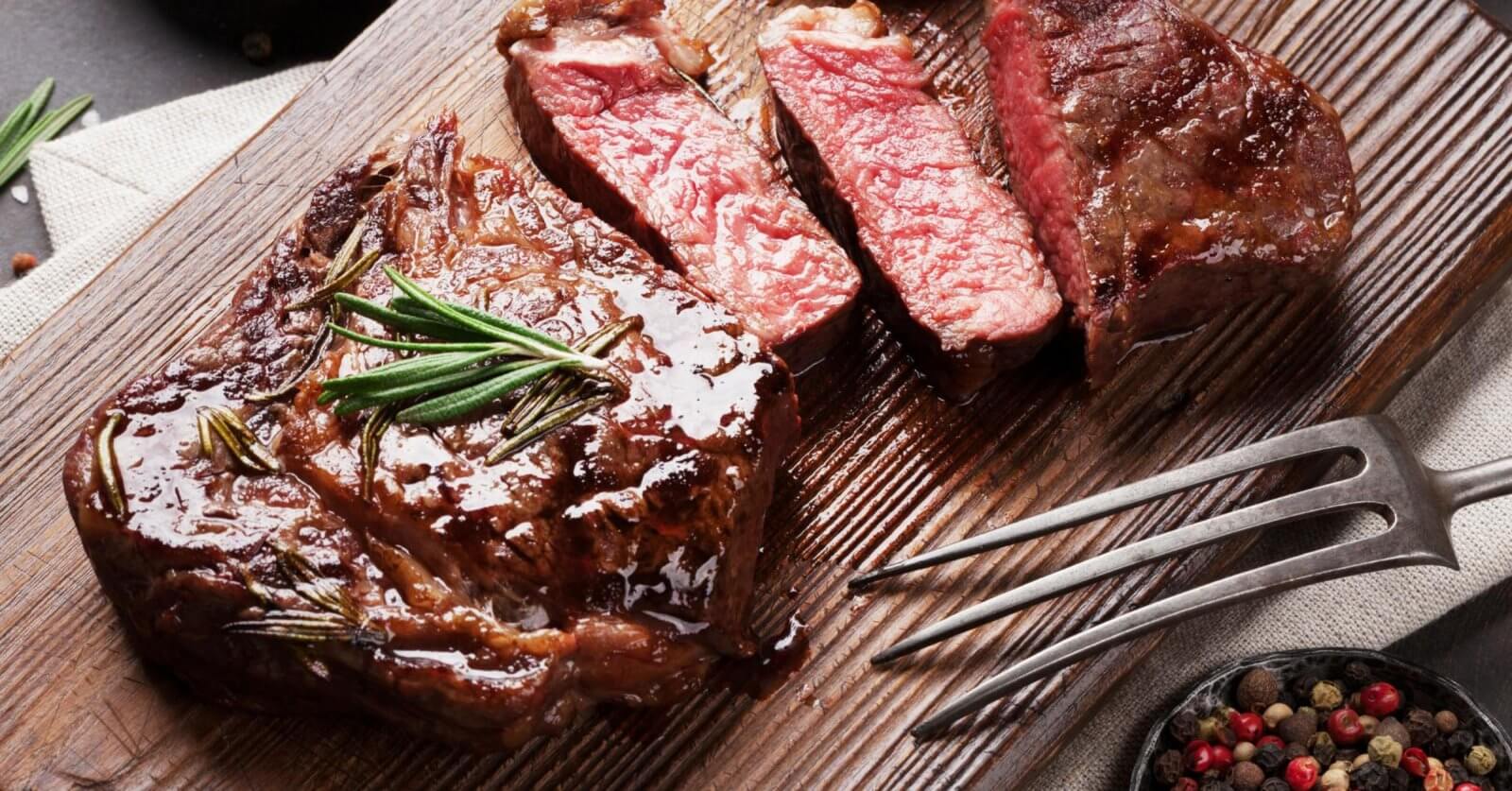 A wooden cutting board with a juicy, medium-rare steak is garnished with fresh rosemary and sliced into three pieces. A vintage fork rests on the board, beside a small bowl of colorful peppercorns. The neutral-toned cloth backdrop lends a charming vibe, reminiscent of delightful dining spots in Salcedo Village.