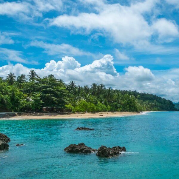 A serene tropical beach with clear turquoise waters, scattered rocks, and lush green vegetation along the shoreline. The sky is bright blue with scattered white clouds. Palm trees and dense forest extend to the edge of the sandy beach at one of the Best Samal Islands Resorts, creating a picturesque coastal landscape.