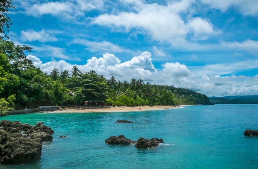 A serene tropical beach with clear turquoise waters, scattered rocks, and lush green vegetation along the shoreline. The sky is bright blue with scattered white clouds. Palm trees and dense forest extend to the edge of the sandy beach at one of the Best Samal Islands Resorts, creating a picturesque coastal landscape.