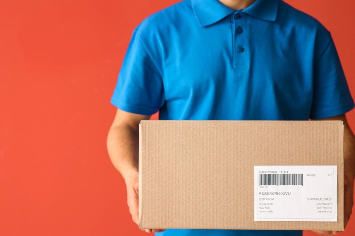 A person in a blue polo shirt holds a cardboard box with a shipping label, likely ready for delivery to a Shopee Xpress Drop-off Point. Set against a solid orange-red background, the box is centered in the image, though the text on its label isn’t fully readable. The person's head remains out of view.