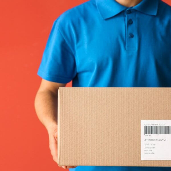 A person in a blue polo shirt holds a cardboard box with a shipping label, likely ready for delivery to a Shopee Xpress Drop-off Point. Set against a solid orange-red background, the box is centered in the image, though the text on its label isn’t fully readable. The person's head remains out of view.