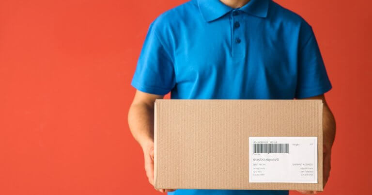 A person in a blue polo shirt holds a cardboard box with a shipping label, likely ready for delivery to a Shopee Xpress Drop-off Point. Set against a solid orange-red background, the box is centered in the image, though the text on its label isn’t fully readable. The person's head remains out of view.
