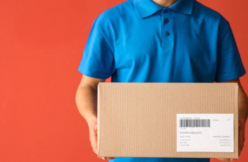 A person in a blue polo shirt holds a cardboard box with a shipping label, likely ready for delivery to a Shopee Xpress Drop-off Point. Set against a solid orange-red background, the box is centered in the image, though the text on its label isn’t fully readable. The person's head remains out of view.