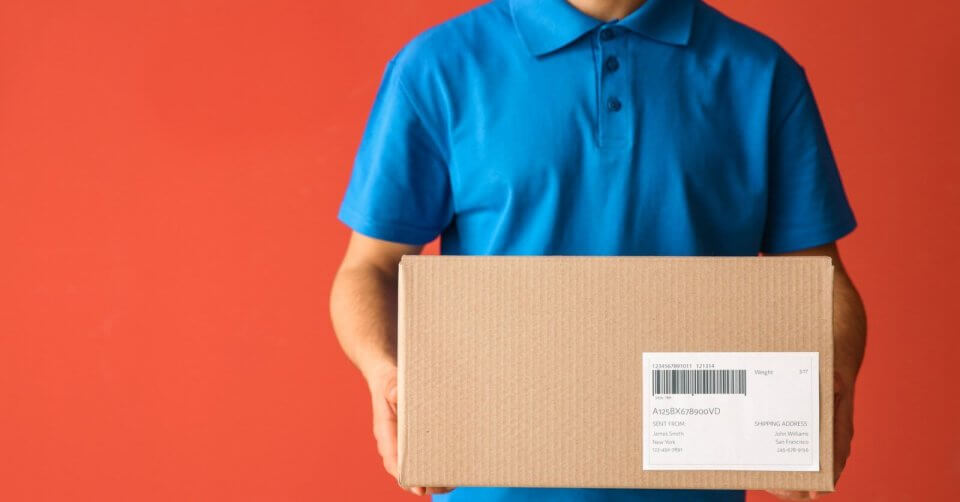 A person in a blue polo shirt holds a cardboard box with a shipping label, likely ready for delivery to a Shopee Xpress Drop-off Point. Set against a solid orange-red background, the box is centered in the image, though the text on its label isn’t fully readable. The person's head remains out of view.