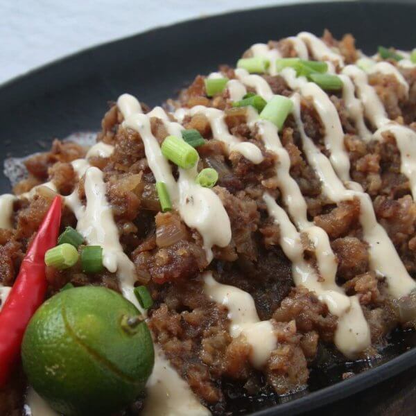 A sizzling platter of sisig is topped with creamy mayonnaise drizzles and garnished with chopped green onions. A whole red chili pepper and a halved calamansi adorn the side, enhancing the dish's vibrant presentation. This culinary delight, perfect to visit at San Fernando restaurants, is set against a neutral background.