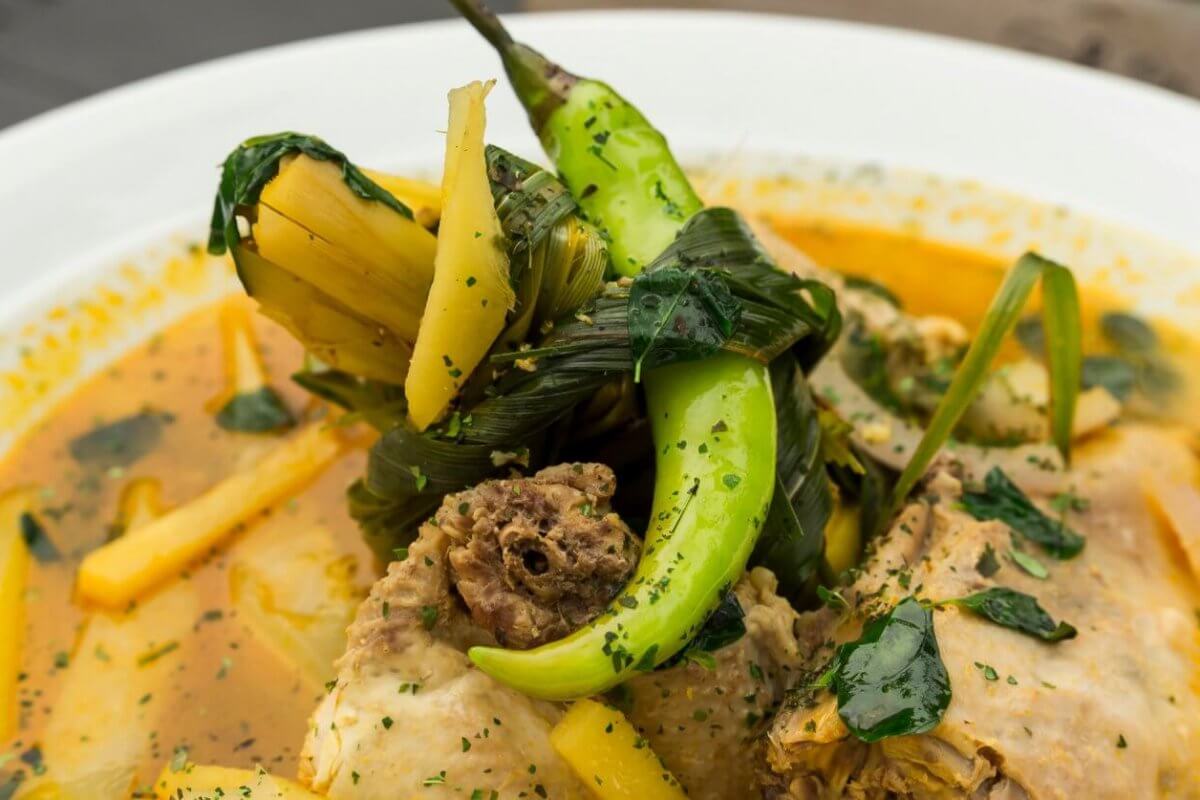 A close-up of a bowl of soup features tender meat pieces, green chili peppers, leafy greens, and yellow vegetable slices in a yellowish broth. The soup is garnished with herbs and served in a white bowl. The richly seasoned broth delivers an aromatic experience you'd expect from one of SM Megamall's best restaurants.