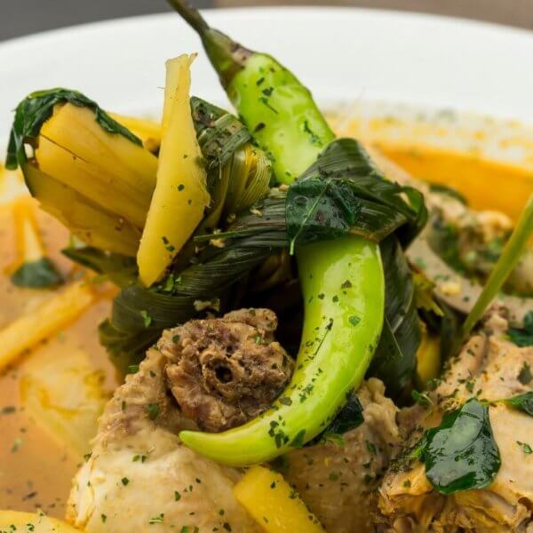 A close-up of a bowl of soup features tender meat pieces, green chili peppers, leafy greens, and yellow vegetable slices in a yellowish broth. The soup is garnished with herbs and served in a white bowl. The richly seasoned broth delivers an aromatic experience you'd expect from one of SM Megamall's best restaurants.