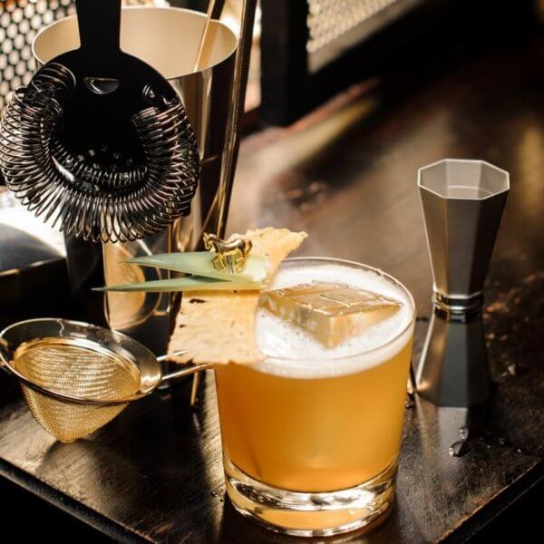 A well-lit bar setup in a hidden gem of Manila features a cocktail garnished with a pineapple leaf and dried pineapple slice on a wooden surface. Nearby are a strainer, shaker, jigger, and fine mesh sieve, evoking the professional mixology found in speakeasy bars.