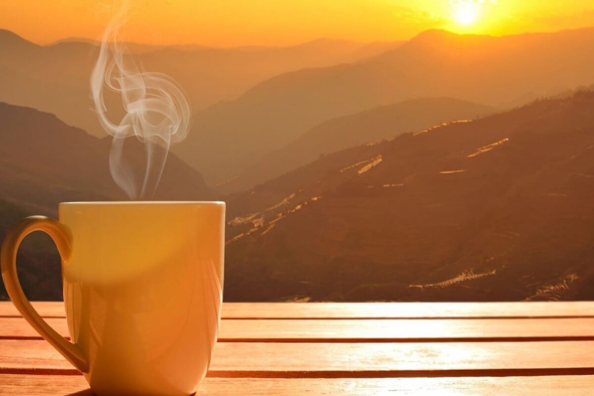 A steaming mug sits on a wooden surface, with a breathtaking sunrise over the mountain ranges of Tanay, Rizal in the background. The sky displays hues of orange and yellow, casting a warm glow over the landscape. Mist gently rises from the valleys, adding a serene and tranquil atmosphere to one of the best coffee spots you'll ever find.