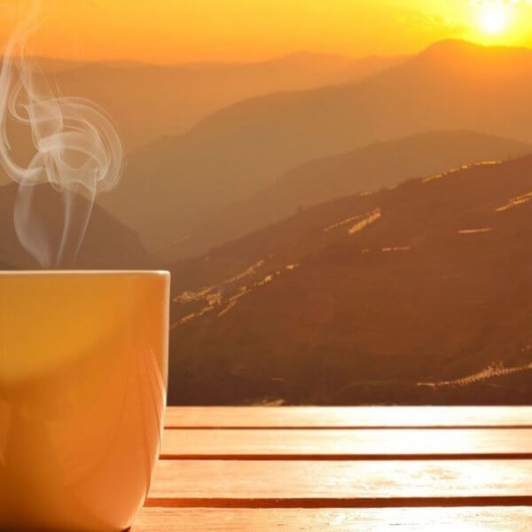 A steaming mug sits on a wooden surface, with a breathtaking sunrise over the mountain ranges of Tanay, Rizal in the background. The sky displays hues of orange and yellow, casting a warm glow over the landscape. Mist gently rises from the valleys, adding a serene and tranquil atmosphere to one of the best coffee spots you'll ever find.