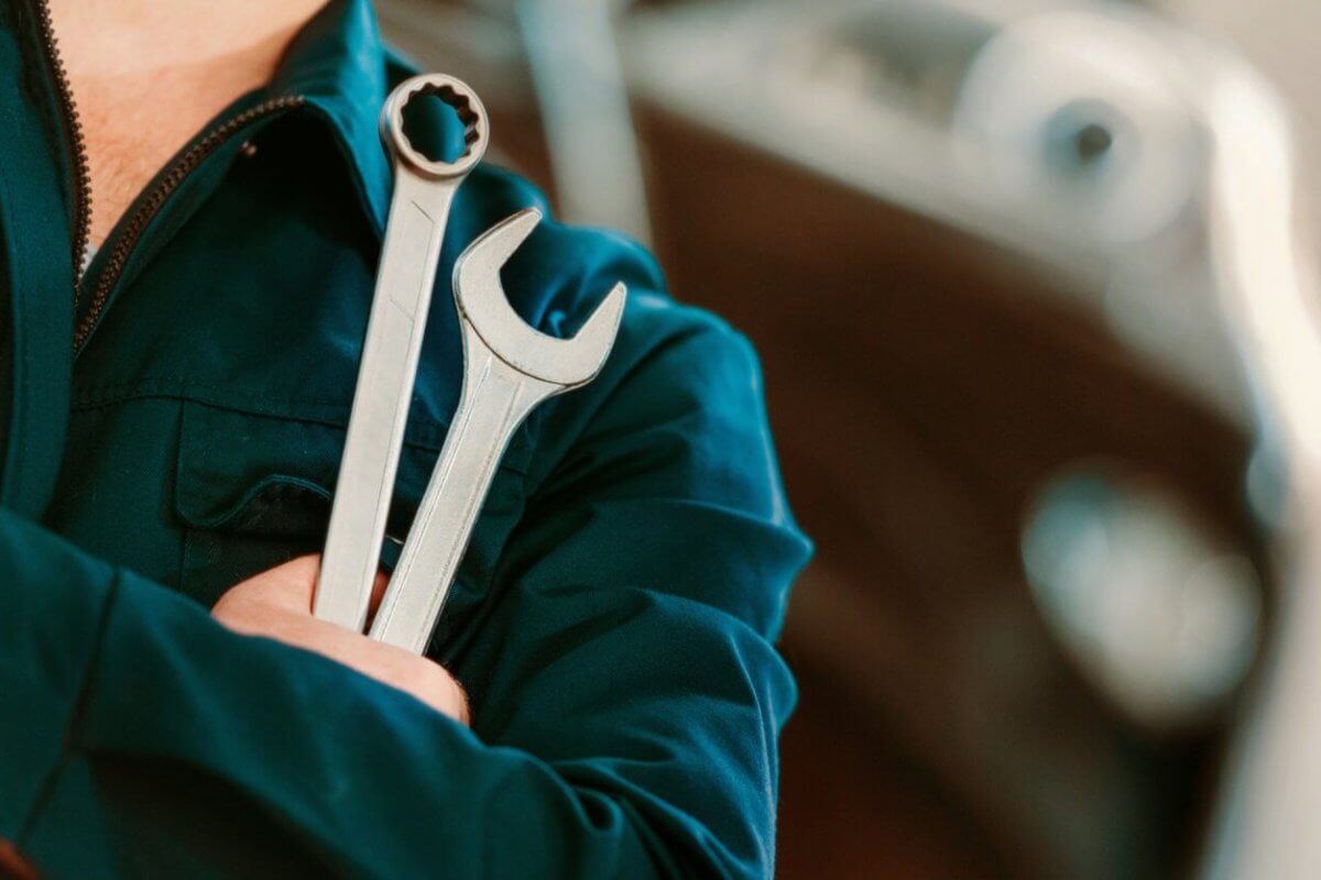 A person in a dark blue mechanic's uniform stands with arms crossed, holding two wrenches. One wrench has an open-end head, and the other has a box-end head. The background is blurred, showing the contours of a vehicle, suggesting an automotive repair setting at one of the top TESDA Accredited Competency Assessment Centers.