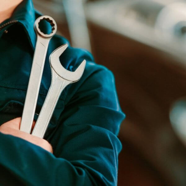 A person in a dark blue mechanic's uniform stands with arms crossed, holding two wrenches. One wrench has an open-end head, and the other has a box-end head. The background is blurred, showing the contours of a vehicle, suggesting an automotive repair setting at one of the top TESDA Accredited Competency Assessment Centers.
