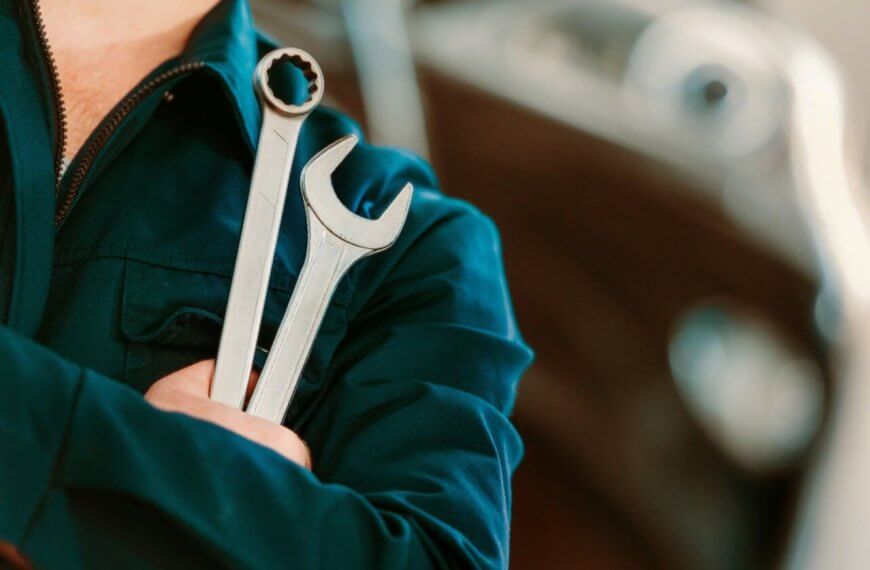 A person in a dark blue mechanic's uniform stands with arms crossed, holding two wrenches. One wrench has an open-end head, and the other has a box-end head. The background is blurred, showing the contours of a vehicle, suggesting an automotive repair setting at one of the top TESDA Accredited Competency Assessment Centers.