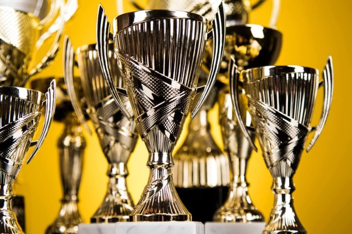 A collection of shiny silver trophies with intricate designs, featuring two handles each and mounted on marble bases, are displayed closely together. The background is a solid yellow color, emphasizing the polished surfaces and reflective qualities of these prestigious awards from top trophy suppliers.